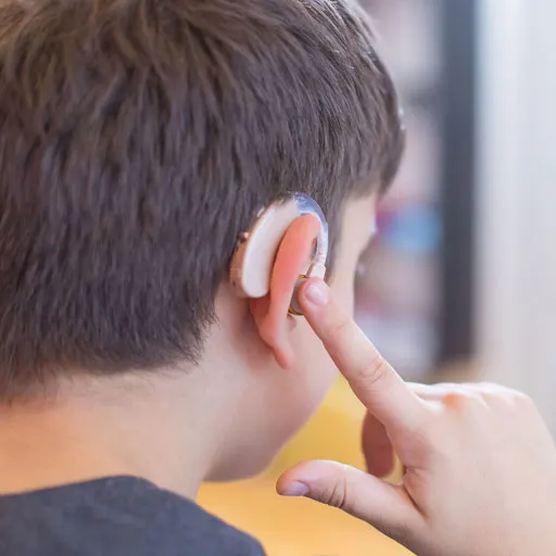 Young boy wearing a skin-colored hearing aid; he's tapping it to adjust or control sound settings.